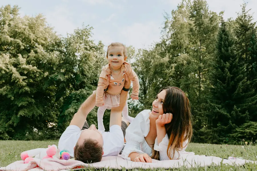 Tata podrzuca córeczkę na kocu w parku, mama się śmieje